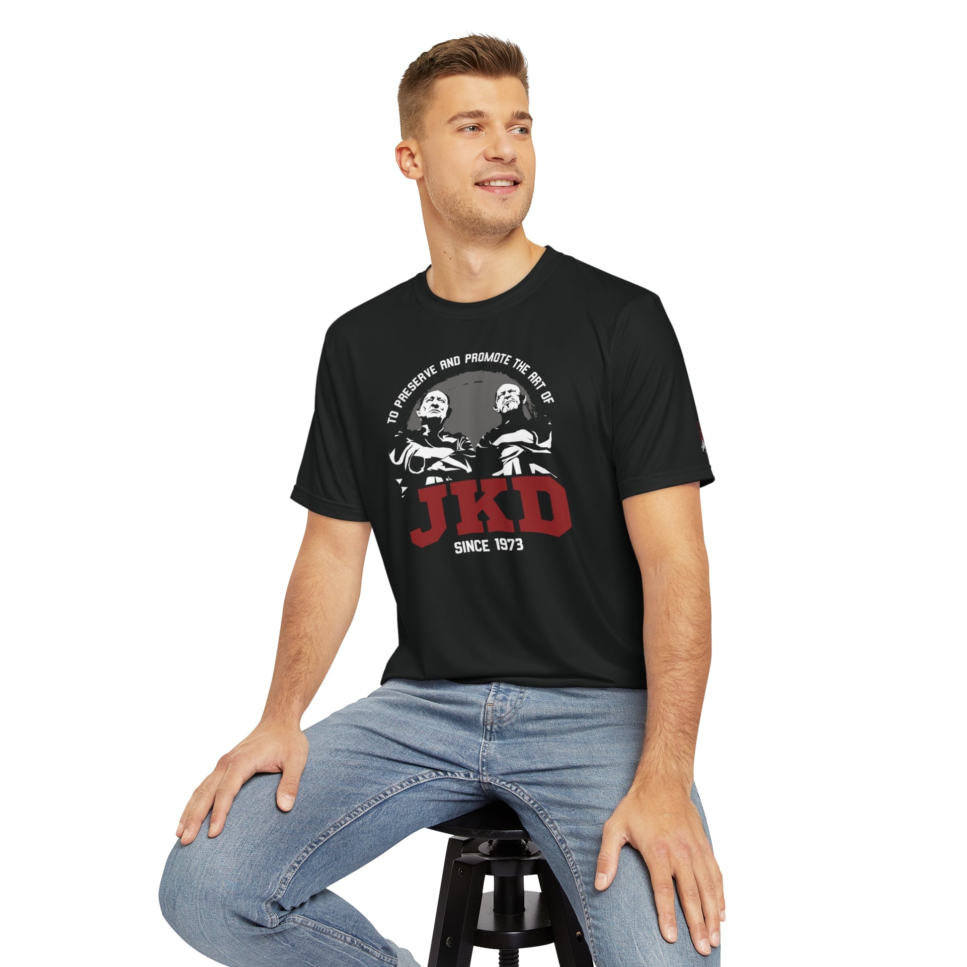 Front view of a man sitting on a stool, wearing a black polyester t-shirt with an image of Tim Tackett and Bob Bremer, two prominent Jeet Kune Do teachers. The text surrounding the image reads "To preserve and promote the art of JKD".