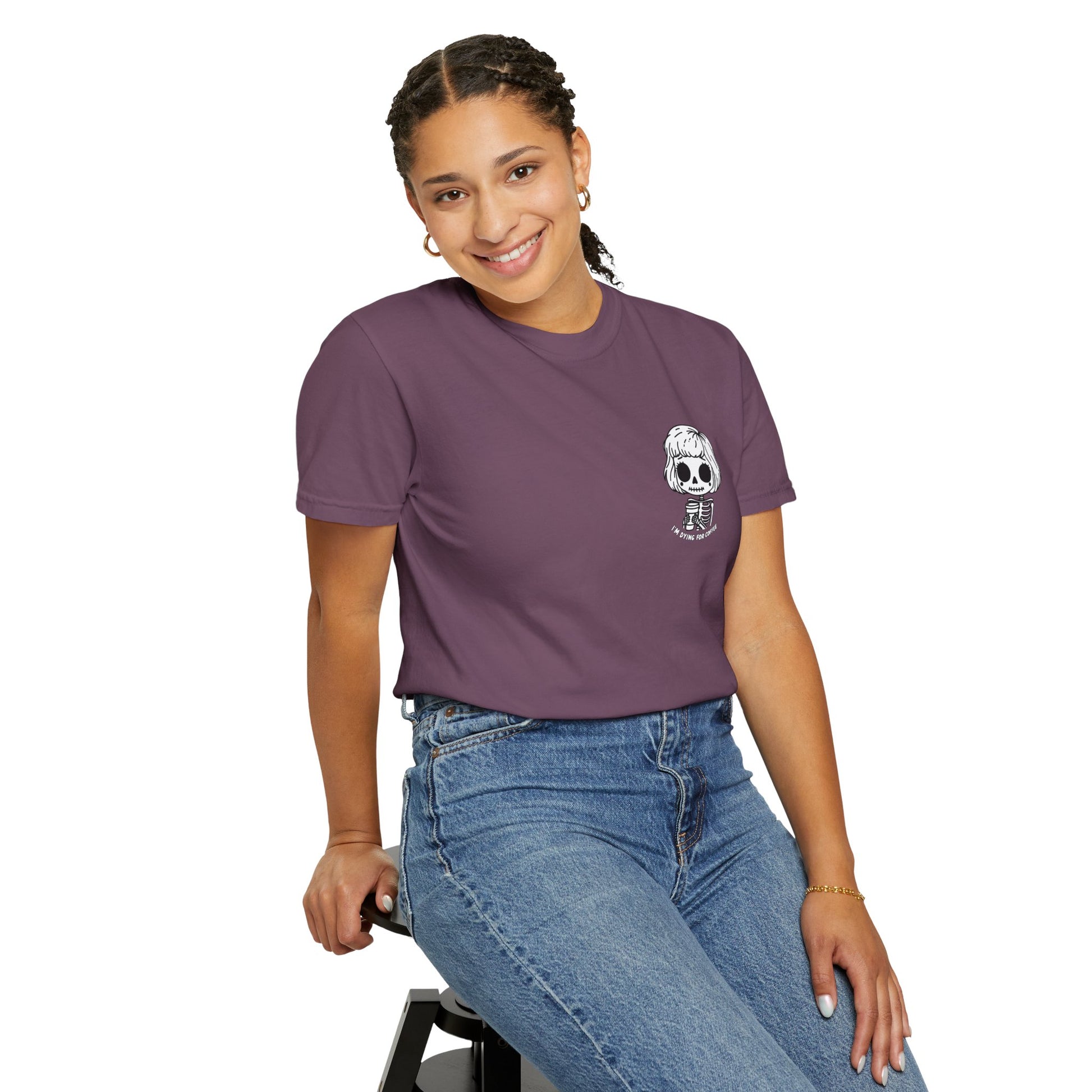 Woman seated on a stool, wearing a berry colored t-shirt with a design of a skeleton girl holding a cup of coffee. Written underneath is the text "I'm dying for coffee". The design is placed on the left chest area of the t-shirt.