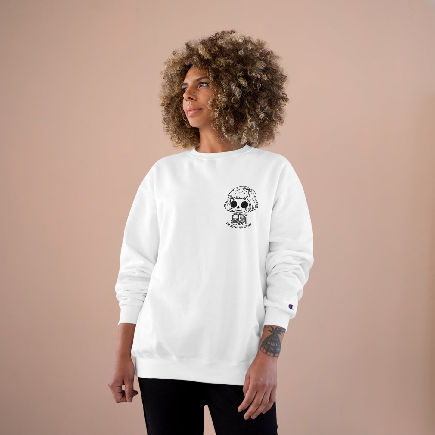 Woman wearing a white sweatshirt with a design of a skeleton girl holding a cup of coffee. Written underneath is the text "I'm dying for coffee". The design is placed on the left chest area of the sweatshirt.