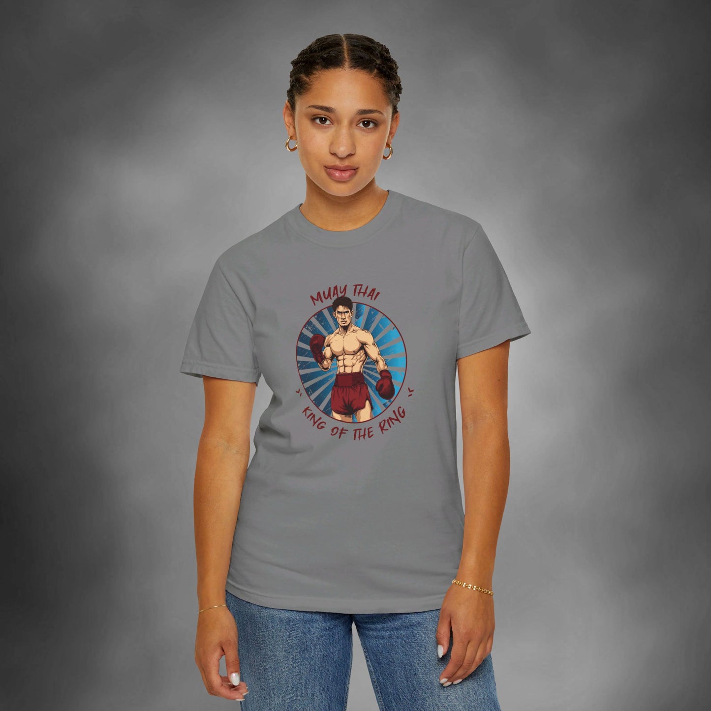 A woman wearing a gray, Comfort Colors cotton t-shirt, with an illustration of a Thai boxer in the center of the chest. Written around the illustration is the text, "Muay Thai - King of the Ring".