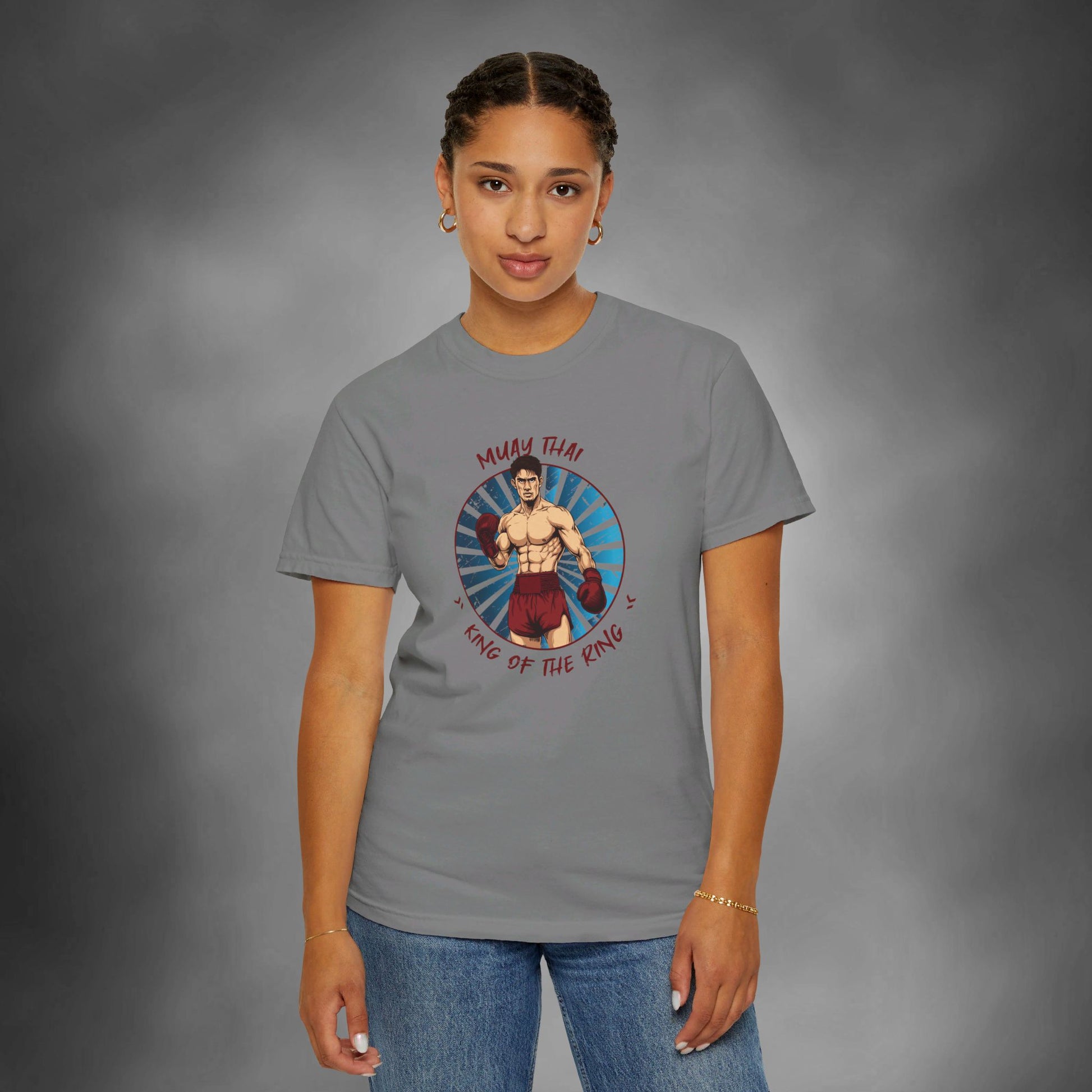 A woman wearing a gray, Comfort Colors cotton t-shirt, with an illustration of a Thai boxer in the center of the chest. Written around the illustration is the text, "Muay Thai - King of the Ring".