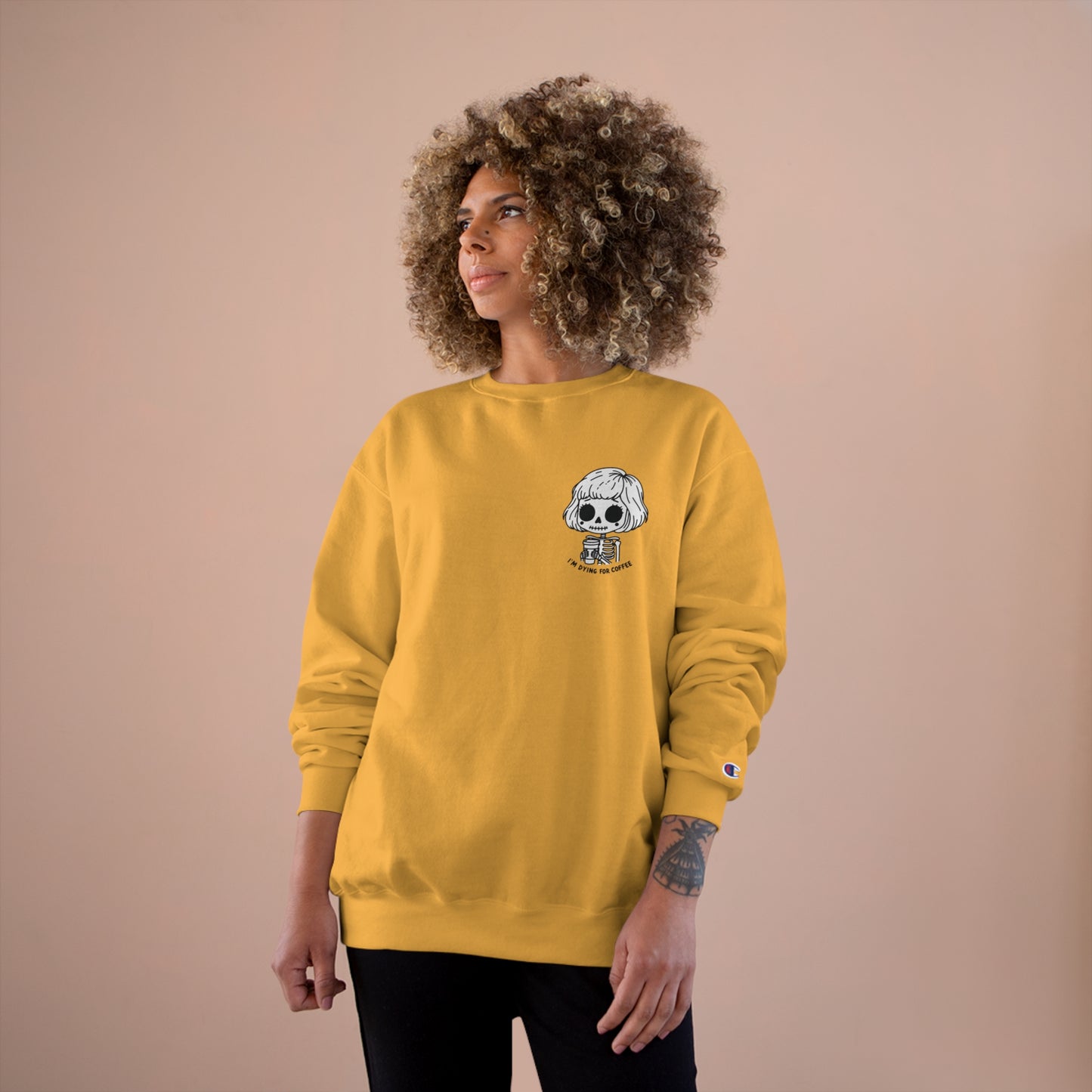 Woman wearing a gold colored sweatshirt with a design of a skeleton girl holding a cup of coffee. Written underneath is the text "I'm dying for coffee". The design is placed on the left chest area of the sweatshirt.