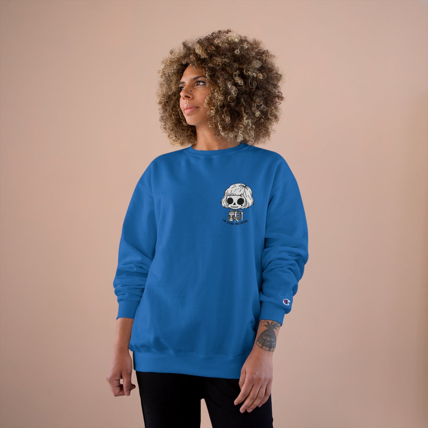 Woman wearing a royal blue colored sweatshirt with a design of a skeleton girl holding a cup of coffee. Written underneath is the text "I'm dying for coffee". The design is placed on the left chest area of the sweatshirt.