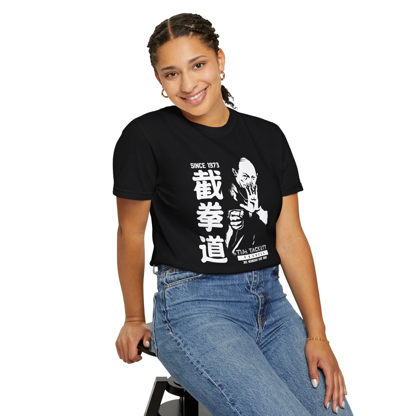 Woman, seated on a stool, wearing a black cotton t-shirt with an illustration of martial arts instructor Tim Tackett on the front. The text surrounding the design reads "Jeet Kune Do since 1973".