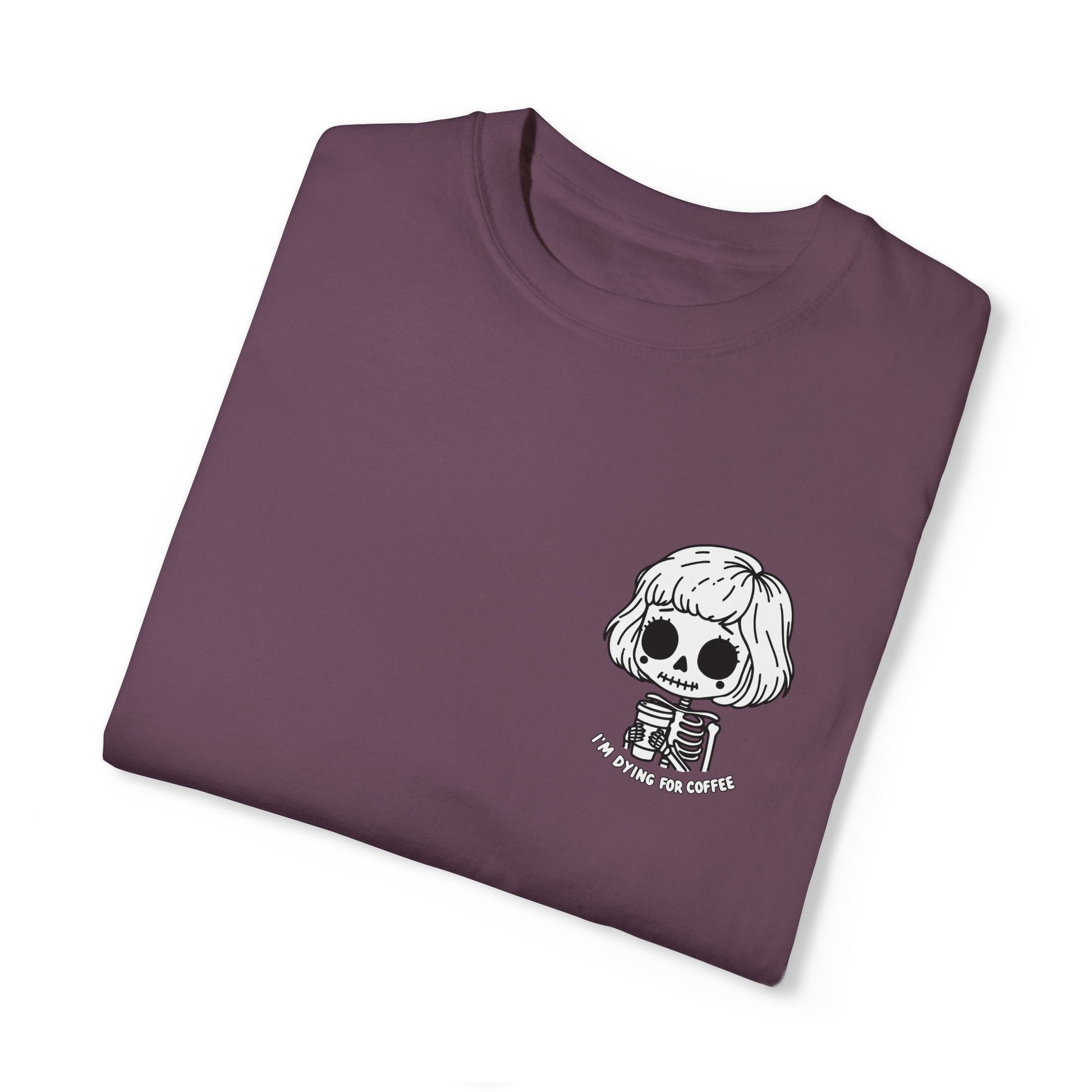 A folded,  berry colored t-shirt with a design of a skeleton girl holding a cup of coffee. Written underneath is the text, "I'm dying for coffee". The design is placed on the left chest area of the t-shirt.