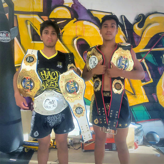 Muay Thai and MMA fighters Adam and Noah Penunuri, with multiple championship belts draped over their shoulders. The man on the left, Adam Penunuri, is wearing a custom fight team jersey from MartialBoutique.