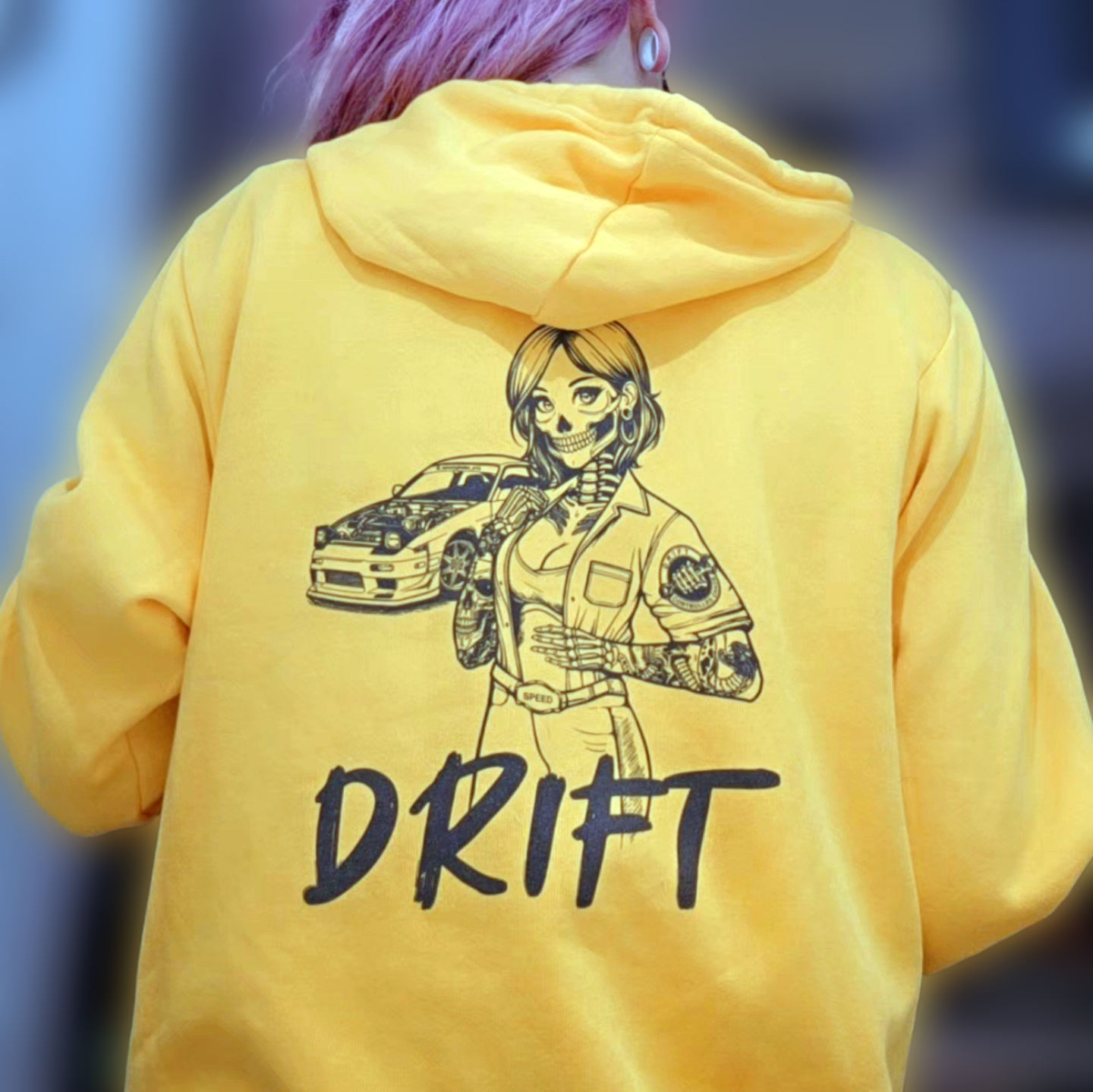 A woman wearing a gold colored fleece hoodie, with a design of a female skeleton dressed as a mechanic on the back. The mechanic is standing next to her car with the word "Drift" written underneath.